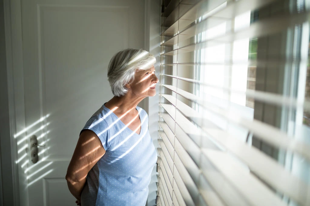older woman looking through blinds on a window